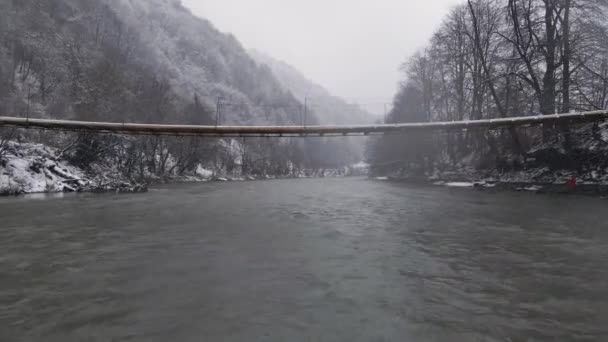 Vista del dron volando hacia adelante sobre el río y debajo del puente de cable en Blizzard clima nevado — Vídeo de stock
