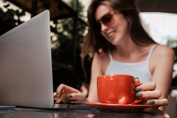 Молодая веселая женщина пьет и работает на ноутбуке в кафе на открытом воздухе, сосредоточиться на чашке кофе, внештатный концепт — стоковое фото