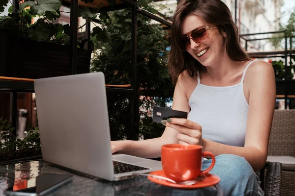 Молодая женщина с ноутбука и проведение кредитных карт, сидя в кафе на открытом воздухе, концепция интернет-магазинов — стоковое фото