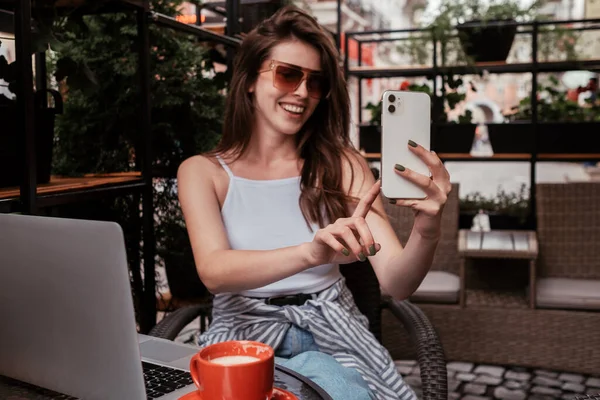 Молодая веселая женщина с помощью смартфона для видеозвонка, сидя в кафе на открытом воздухе — стоковое фото