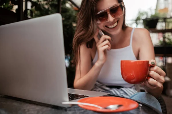 Sorrindo jovem morena mulher falando no telefone celular enquanto sentado com laptop e café em um café ao ar livre — Fotografia de Stock