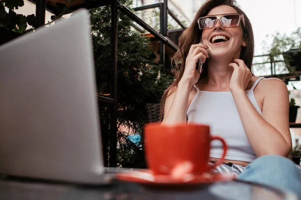 Sorrindo jovem morena mulher falando no telefone celular enquanto sentado com laptop e café em um café ao ar livre — Fotografia de Stock