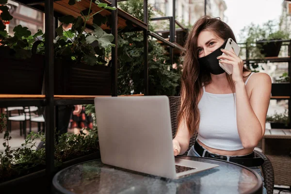 Jovem morena mulher vestindo máscara protetora do rosto falando no telefone móvel enquanto sentado com laptop em um café ao ar livre — Fotografia de Stock