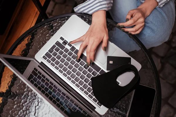 Mujer joven usando el ordenador portátil mientras está sentado en un café al aire libre, tarjeta de crédito y máscara facial protectora en el cuaderno, Covid-19 Concepto de compras en línea —  Fotos de Stock