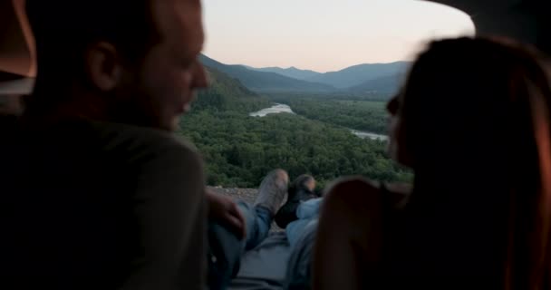 車のトランクに寝そべって、地平線でロードトリップ、山と川を楽しむ幸せな若いカップルの後部ビュー — ストック動画