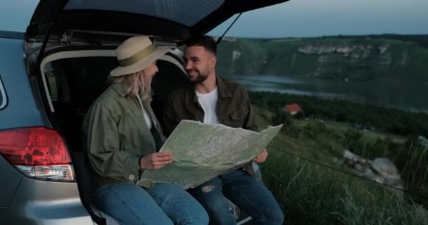 Homem e mulher sentados no tronco do carro, usando o mapa de papel na viagem de fim de semana, viagem e conceito de aventura — Vídeo de Stock