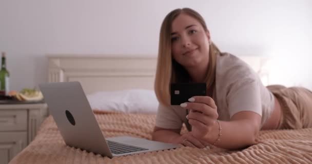 ホテルの部屋のベッドに横になり、カメラにクレジットカードを表示する若いブロンドの女性,ショッピングオンライン — ストック動画