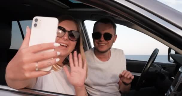 Молодой человек и женщина сидят в машине с видом на море из окна и используют смартфон для видеозвонков, машут руками, чтобы поздороваться во время поездки — стоковое видео