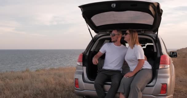 Joven y mujer divirtiéndose en viaje por carretera, sentados en el baúl abierto de su coche todoterreno con vista al mar al borde de la tierra — Vídeo de stock