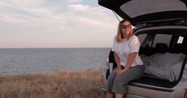 Ung kvinna som har roligt på väg resa, sitter i den öppnade bagageluckan i Suv Car med havsutsikt vid kanten av land — Stockvideo