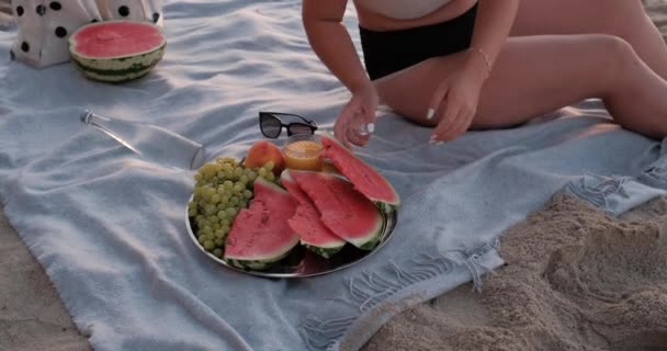 Veduta del picnic con molti frutti e bottiglia d'acqua sulla spiaggia di sabbia del mare prima dell'alba, delle vacanze e del concetto di fine settimana, la ragazza in cappello prende fetta di anguria e sorride alla macchina fotografica — Video Stock