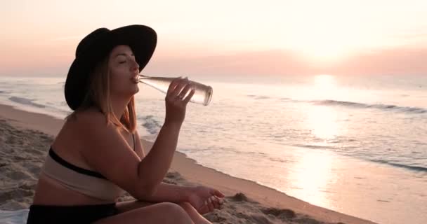 Vista lateral de una mujer joven vestida traje de baño y sombrero agua potable de botella de vidrio en la playa del mar de arena al amanecer — Vídeo de stock