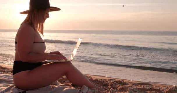 Närbild Side View of Young Woman Klädd baddräkt och hatt Sitter på Sandy Sea Beach vid soluppgången gryningen och arbetar på laptop, frilans och fjärrarbete koncept — Stockvideo