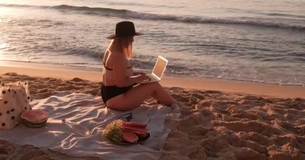 Vista lateral del traje de baño y el sombrero vestidos de mujer joven sentados en la playa del mar de arena al amanecer y trabajando en el concepto de trabajo portátil, independiente y remoto — Vídeo de stock