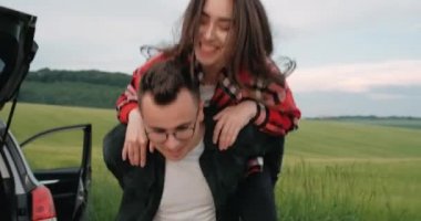 Yolculukta eğlenen genç çift, kırsal kesimde arabalarının yanında sevgilisinin sırtına atlıyor.