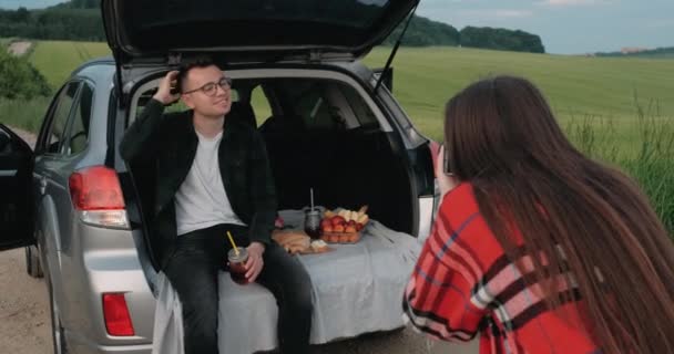 Guy sentado no carro tronco enquanto sua namorada tira uma foto dele em uma câmera de filme antigo, jovens desfrutando de viagem de carro através do campo — Vídeo de Stock