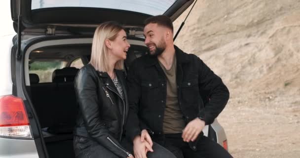 Jong paar, Man en vrouw gekleed Alike in Black Jacket, Veel plezier tijdens het zitten in de geopende kofferbak van de auto, Lachen en lachen tijdens Road Trip — Stockvideo