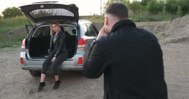 Joven fotografiando a la novia que está sentada en el baúl abierto del coche, chica delante de la cámara — Vídeos de Stock