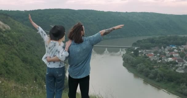 Pareja joven y elegante, hombre y mujer tatuados con sombrero vestidos al estilo boho, abrazando y levantando las manos mientras están de pie al borde del río Canyon — Vídeos de Stock