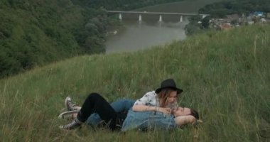 Genç Tarz sahibi Çift, Uzun Saçlı Adam ve Şapkalı Kadın Boho Stili Giyinen, Tepenin Üzerinde Çimenlerde Uzanıyorken Eğlenen, Arka planda Kanyon Nehrinde, Kamera Modellere İlerliyor