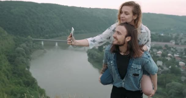 Mladý stylový pár, žena oblečená v Boho stylu sedí na zádech dlouhosrstého muže a přičemž Selfie na Smartphone, zatímco stojí na okraji kaňonu řeky v deštivém letním dni — Stock video