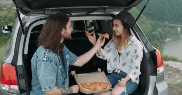 Два молодых путешественника едят пиццу в багажнике автомобиля, стильный мужчина и женщина, одетые в стиле Бохо Наслаждаясь поездкой, концепция путешествия — стоковое видео