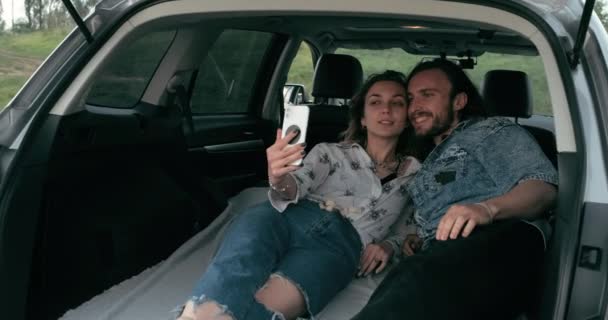 Dois jovens viajantes levando selfie no smartphone dentro do tronco do carro, homem e mulher vestidos em estilo boho desfrutando de viagem juntos, viagem e conceito de férias — Vídeo de Stock