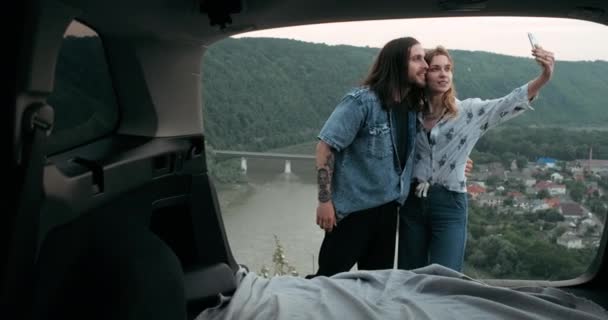 Vista posterior desde el interior del coche de dos jóvenes viajeros que se toman selfie en el teléfono inteligente contra el fondo panorámico, el hombre y la mujer en el estilo Boho disfrutando de un viaje por carretera juntos, viajes y concepto de vacaciones — Vídeo de stock