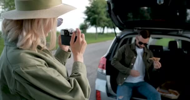 Młoda kobieta robi zdjęcia swojego chłopaka jedzącego pizzę w bagażniku samochodu — Wideo stockowe