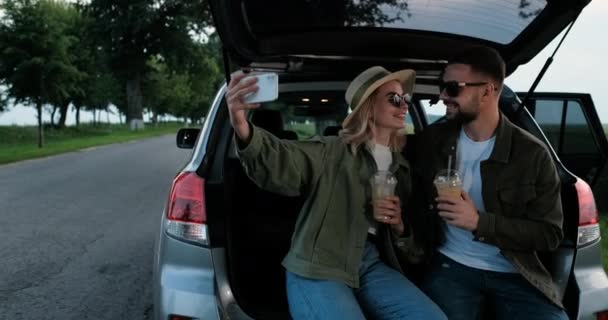 Jonge man en vrouw gekleed Alike, Zittend in de kofferbak van hun auto en drinken koude koffie, Met behulp van Smartphone voor Selfie — Stockvideo