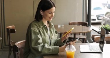 Khaki Ceketli Esmer Kadın, Akıllı Telefon ve Kredi Kartıyla Terrace Cafe 'de Çevrimiçi Ödeme Yapıyor, Masa, Serbest ve Uzaktan Çalışma Konseptinde Taze Meyve Suyu ve Laptop