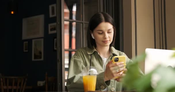 Barna nő Khaki dzseki Nézz körül mosollyal Használata közben okostelefon és fülhallgató, miközben dolgozik Laptop a Terrace Cafe, Friss gyümölcslé az asztalon, Szabadúszó és távmunka koncepció — Stock videók