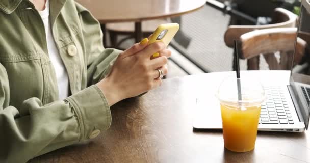 Side View of a Brunette Woman in Khaki Jacket Med Smartphone och hörlurar När du arbetar på laptop på Terrace Cafe, Färsk juice på ett bord, Kameran flyttar från händer till modell ansikte, Freelance Concept — Stockvideo