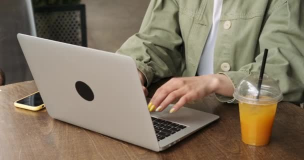 Vista de cerca de una mujer morena en chaqueta caqui usando computadora portátil y auriculares mientras trabajaba en línea en Terrace Café, bebiendo jugo fresco — Vídeo de stock