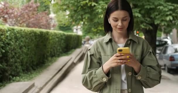 Mladá bruneta žena v khaki bundě pomocí Smartphone a sluchátka pro zatímco procházky venku na ulici na slunné letní den — Stock video