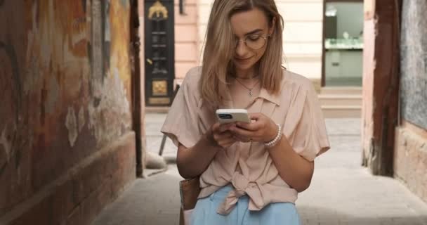 Närbild framifrån av stilig ung kvinna med glasögon och blont hår upptagen skriva på Smartphone medan promenader utomhus på City Street — Stockvideo