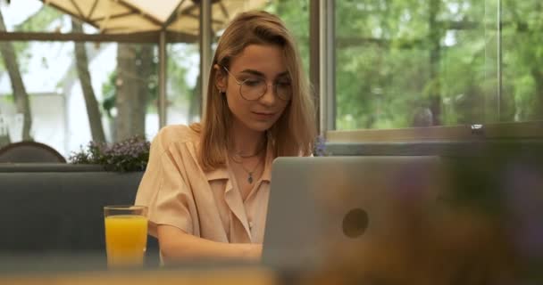 Vista frontal de la joven rubia ocupada trabajando en una computadora portátil y bebiendo jugo de naranja fresco y sonriendo mientras está sentada en el café, freelancer en el trabajo — Vídeos de Stock