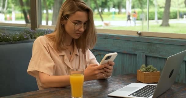 Mulher elegante jovem com óculos e cabelo loiro usando Smartphone e sorrindo enquanto sentado no café no trabalho, laptop e suco de laranja fresco na mesa, Freelance e conceito de trabalho remoto — Vídeo de Stock