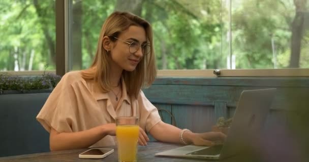 Młoda blondynka w okularach za pomocą laptopa do rozmowy wideo podczas siedzenia w kawiarni, świeży sok pomarańczowy na stole — Wideo stockowe