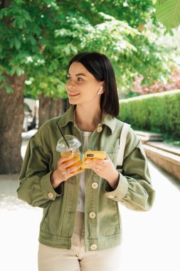 Şehir parkında yürürken akıllı telefonunu ve kulaklıklarını kullanan genç kadın, esmer kız cep telefonu kullanıyor, kokteyl içiyor ve uzaklara bakıyor..