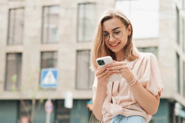 Gözlüklü Neşeli Kadın Akıllı Telefonla Mesaj Yazıyor, Gülümsüyor, Şık Sarışın Kız Açık Hava 'da Cep Telefonuna Bakıyor.