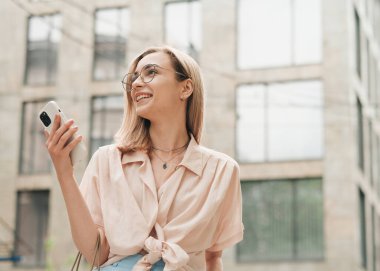 Gözlüklü Genç Kadın Elinde Akıllı Telefonla, Gülümseyen, Zarif Sarışın Kız Sokakta Uzaklara Bakıyor.