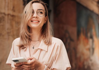 Gözlüklü Neşeli Genç Kadın Portresi Şehirde Yürürken Smartphone 'da mesajlaşıyor, Gözlüklü Sarışın Kız Cep Telefonu Kullanıyor ve Gülümsüyor.