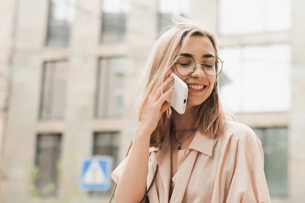 Веселая элегантная женщина в очках с помощью смартфона и улыбки с закрытыми глазами, стильная блондинка на улице, разговаривая через мобильный телефон. — стоковое фото