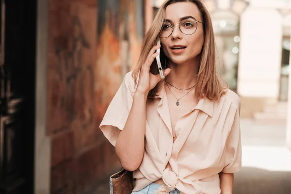 Уверенная в себе элегантная женщина в очках с помощью смартфона во время прогулки по городу, стильная блондинка стоит на улице, разговаривая через мобильный телефон. — стоковое фото