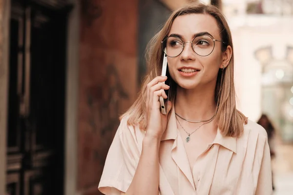 Крупный план веселой элегантной женщины в очках с помощью смартфона и улыбающейся, стильной блондинки в очках, стоящей на улице и разговаривающей через мобильный телефон. — стоковое фото
