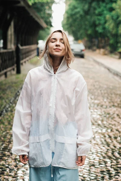 Close Up Ritratto di giovane donna che indossa l'impermeabile mentre in piedi per strada al giorno della pioggia, ragazza bionda vestita in mantello impermeabile divertirsi sotto la pioggia in città. — Foto Stock