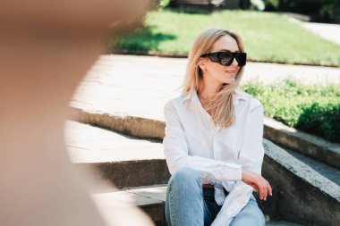 Halka açık parkta merdivenlerde oturan zarif neşeli kadın, güneş gözlüğü takan ve buluşmayı beklerken gülümseyerek uzaklara bakan bayan girişimci..