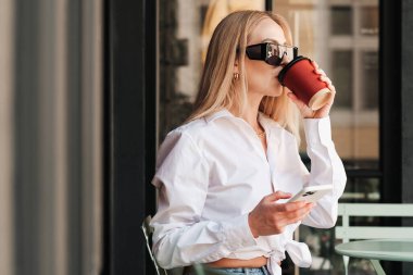 Akıllı Telefon Kullanırken Bir Kafe Masasında Oturan ve İçen Zarif Kadın, Güneş Gözlüğü Giyen ve Beyaz Gömlekli Kadın Kahve Dükkanı Terasında Buluşmayı Bekliyor.