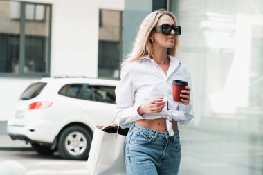 Şık Sarışın Kadın, elinde kahve fincanı olan, kendinden emin, güneş gözlüğü takan ve Altın Mücevher takan, Şehir Caddesinde yürüyen.